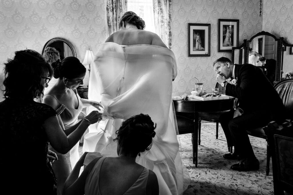 Groom eating a snack while the bride gets help with her dress